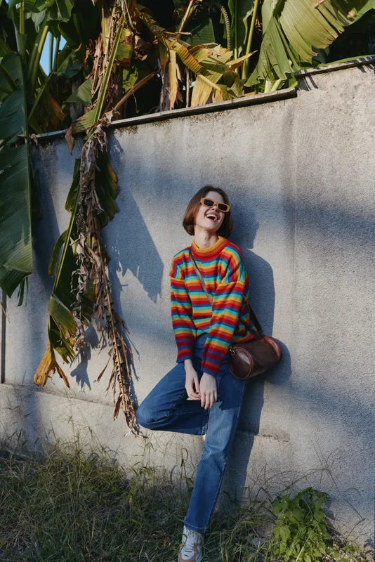 Woman in her mid twenties wearing a colorful striped sweater and sunglasses, smiling while leaning against a concrete wall with tropical leaves, casual outfit, lifestyle travel portrait.