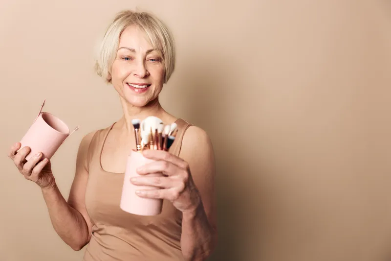 Smiling mature woman holding a pink container with makeup brushes isolated on beige background. Concept of beauty tools, self-care, and joyful lifestyle for older adults.