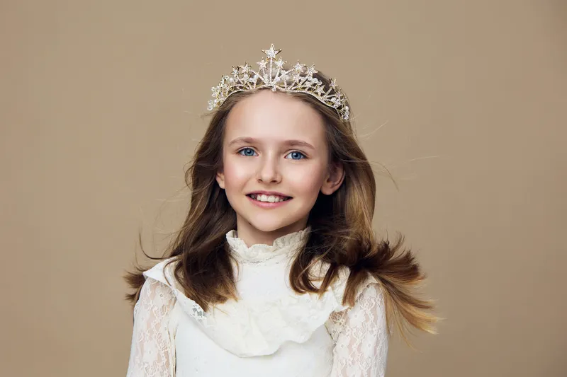 Happy young girl with a crown in elegant dress, showcasing joy and innocence against a soft beige background, perfect for children s fashion themes