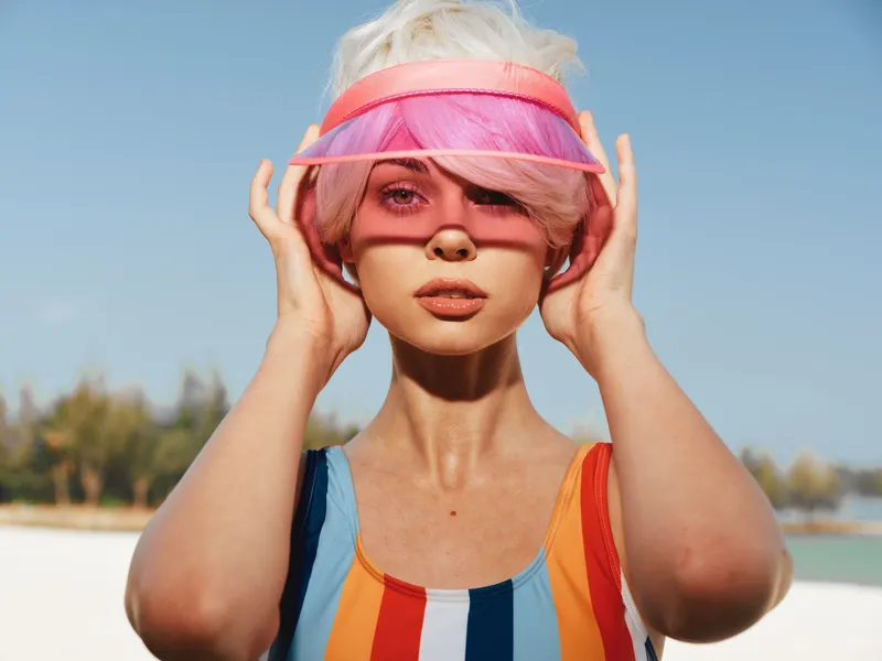Young woman wearing a rainbow striped swimsuit and pink visor stands on a sunny beach, delivering bold colors, confident pose, and a playful summer vibe.