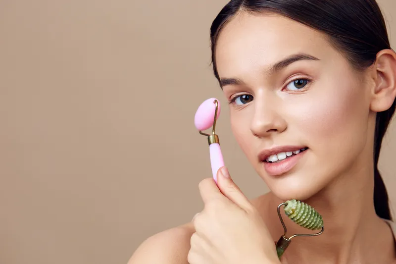 Young woman with natural makeup using facial rollers for skincare, highlighting beauty and wellness concepts, in a soft pastel background