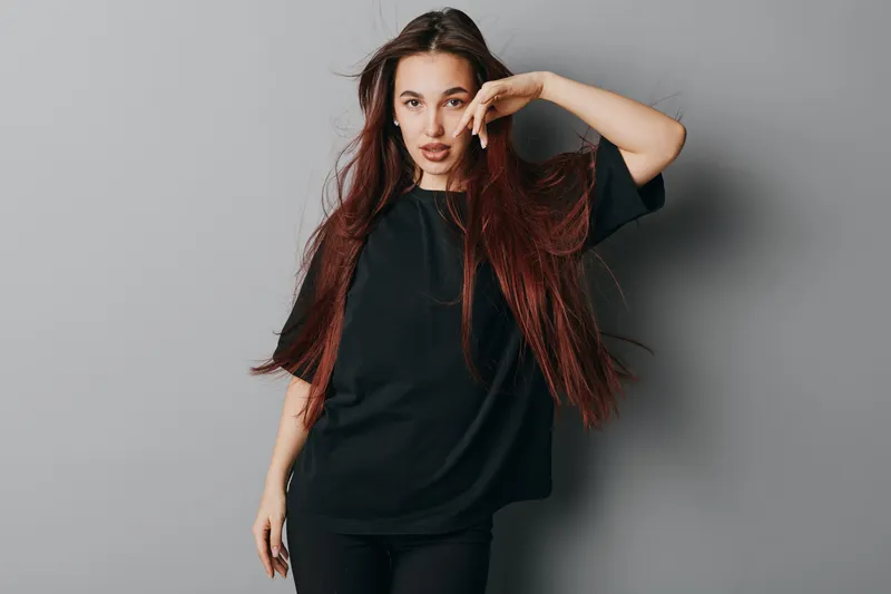 Young woman with long hair wearing black oversized t shirt posing confidently against gray background. Female model standing with hand near face and casual style in studio.