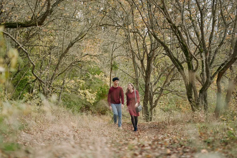 A candid couple walks along a leaf strewn forest path, capturing authenticity and credibility in a natural moment amid autumn light and calm surroundings.