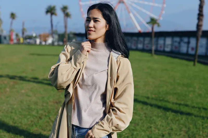 Casual woman outdoors in beige jacket walking on green grass with ferris wheel in distance, sunny day, candid style, everyday fashion, leisure activity, park scenery, relaxed pose, natural light,