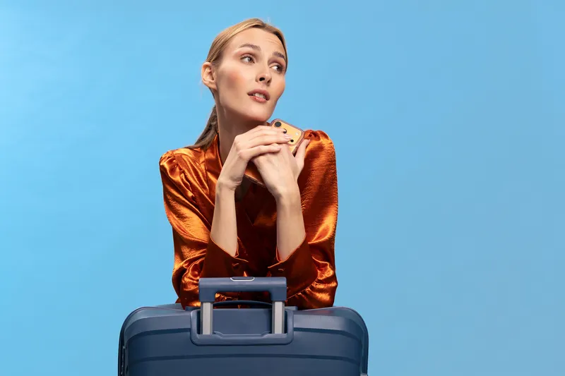Young woman in orange satin blouse holds smartphone and leans on blue suitcase, looking away thoughtfully. Concept of travel, planning, and anticipation in studio with blue background.