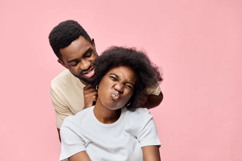 Young black couple having fun, playful expressions, posing together against a soft pink background, casual attire highlighting joy and affection, ideal for love themes