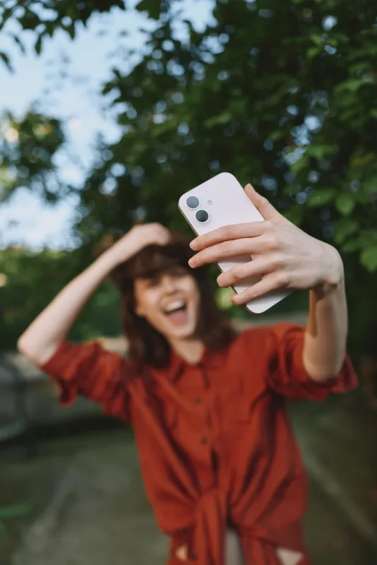 A smiling woman takes a carefree outdoor selfie in a sunny setting, wearing a red dress. The image conveys casual lifestyle, happiness, and self expression