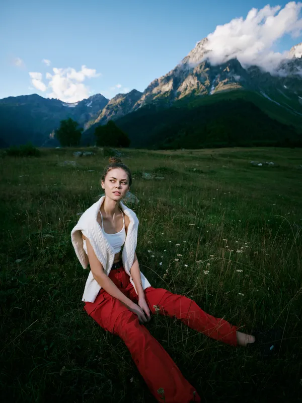 Woman sitting in a meadow with red pants and white top, rugged mountains in the background, cloudy sky, outdoor fashion mood, serene landscape and nature inspired scene