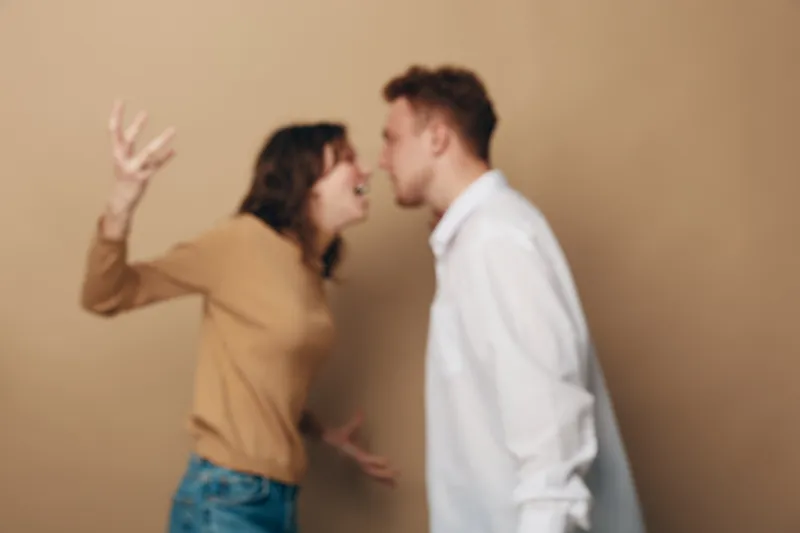 Blurry image of a young man and woman arguing indoors, showing emotional conflict and tension. The woman gestures with raised hand while the man leans forward in disagreement.