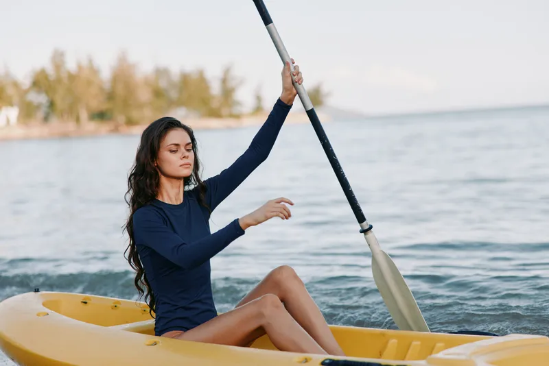 A young woman paddles a yellow kayak across calm blue water, wearing a dark long sleeve top as she rows with a steady stroke, capturing a peaceful outdoor moment.