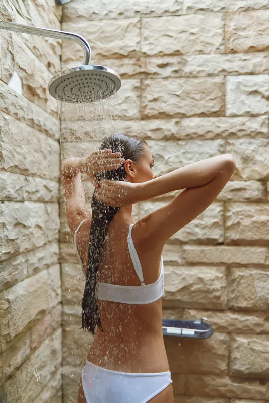 woman showering outdoor with stone wall background, wet hair, natural light, relaxed mood, summer day bathing, healthy skin care routine