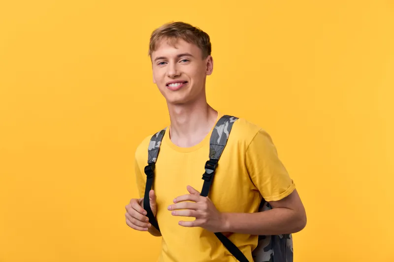 Young man with a backpack smiles against a bright yellow background, expressing casual style, confidence and readiness for travel, school or everyday exploration