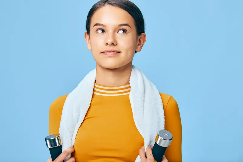 Female athlete in a bright studio, wearing an orange top with a white towel around her neck, holding dumbbells, ready for a workout, showcasing fitness, sport, training, health and active lifestyle.