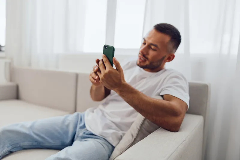 A man using his phone while lounging on a couch, embodying modern anxiety and the blurring lines between work and personal life in a comfortable setting.