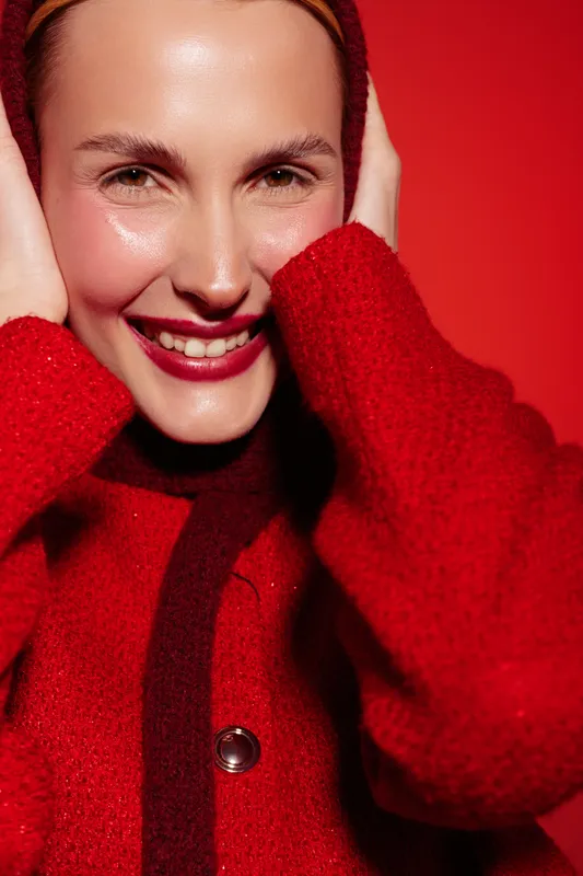 Smiling woman in red sweater poses with hands near face against red background. Close-up portrait with natural makeup and joyful expression. Concept of happiness and fashion.