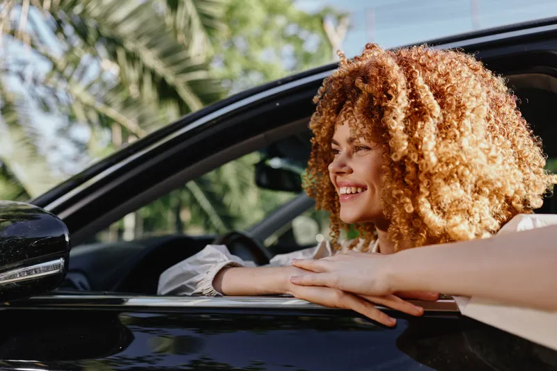 smiling woman with curly hair leaning out of black car window outdoors in bright sunlight, happy, casual, summer vibe, natural background