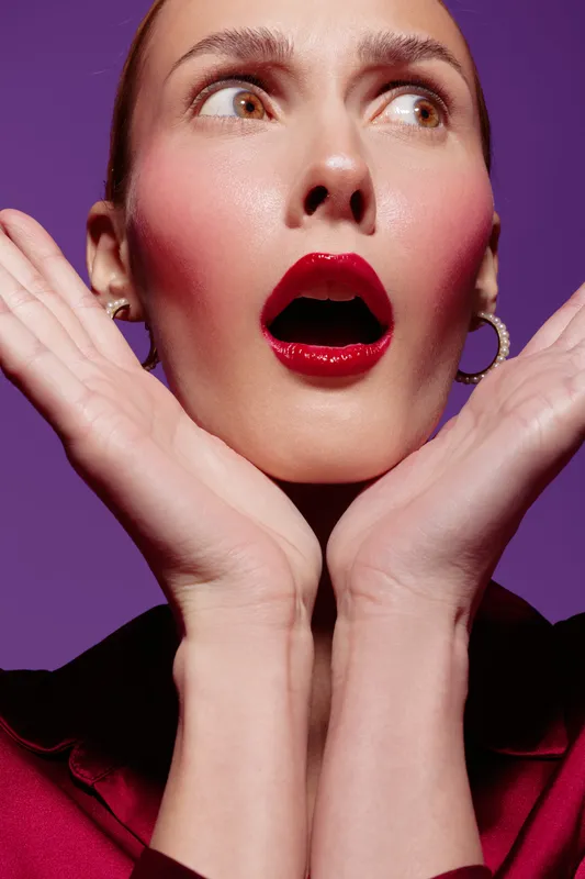 Surprised woman with red lipstick and pink blush holds hands near face on purple background. Concept of emotion, beauty, makeup, and facial expression in studio lighting.