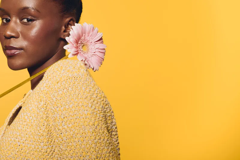 Close-up of a smiling African woman with dark skin wearing a yellow knitted sweater holding a pink flower behind her shoulder on yellow background. Concept of natural beauty and floral elegance.