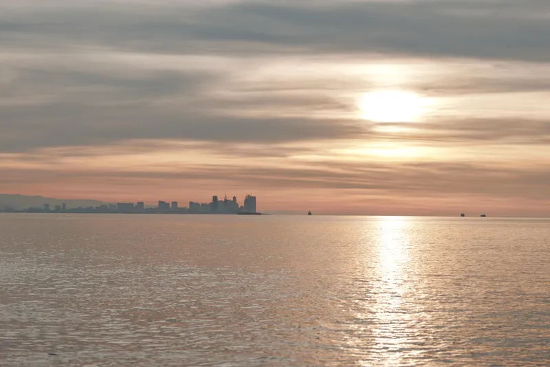 Sunset over calm water with a distant city skyline, warm tones across the sky, and a reflective surface creating a tranquil evening landscape by the shore