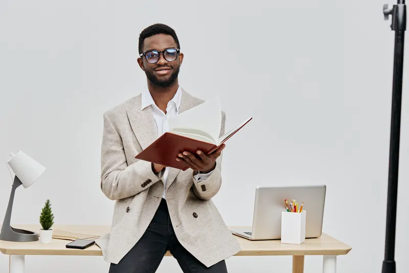 Professional young black man in beige blazer reading a book while standing beside a desk with laptop and stationery, studio portrait, lifestyle and education concept with clean white background.