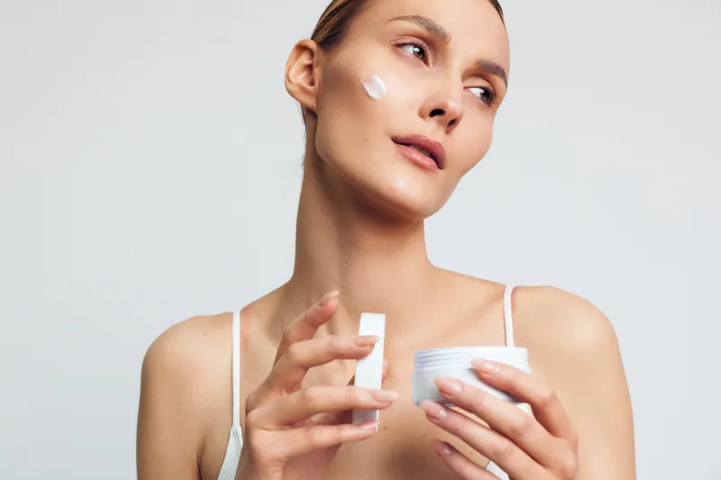 A woman with clear skin applies white cream on her face holding a jar in one hand and a cotton pad in the other. The bright background emphasizes skincare and beauty routine concepts.