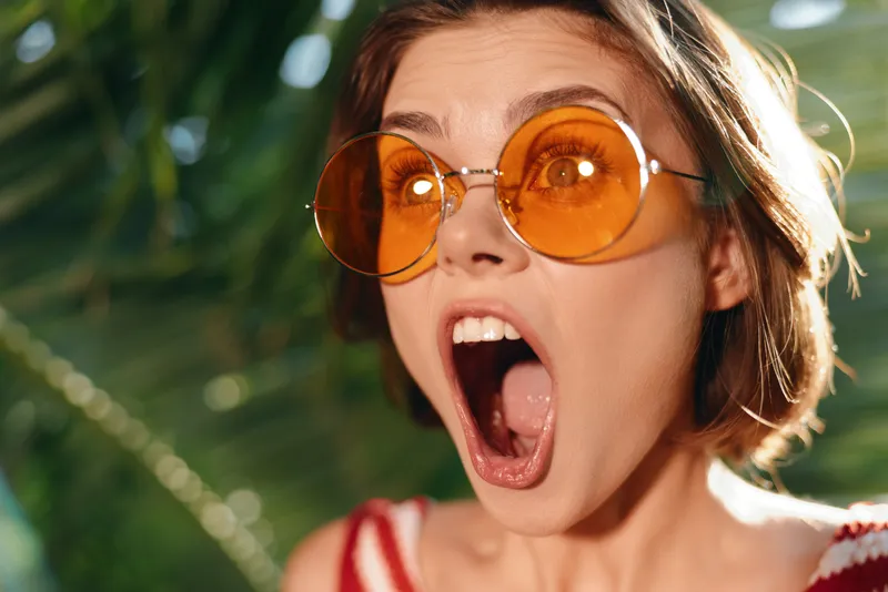 A joyful young woman wearing oversized orange sunglasses in a sunlit tropical setting; she shows surprise with a wide mouth and astonished gaze, creating a vivid, upbeat mood.