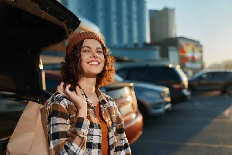 Woman shopping by car trunk smiling in parking lot at golden hour, candid urban portrait conveying authenticity, mindful living and emotional storytelling with warm glow