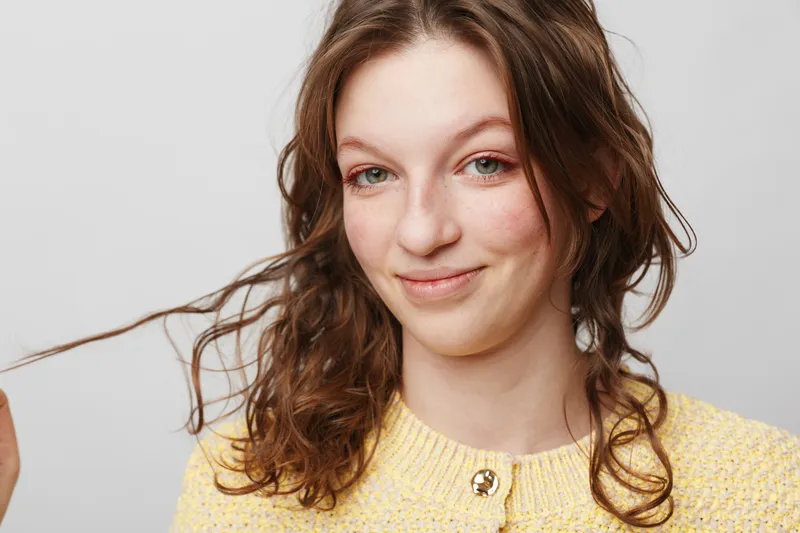 Young woman with curly brown hair smiles gently wearing a yellow sweater against a plain light background. Concept of natural beauty, casual style, and positive emotions.