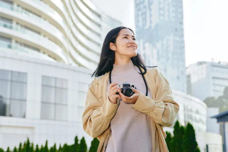 Woman photographer outdoors in an urban setting, holding a camera, casual jacket and soft sweater, exploring city architecture and bright daylight for travel, lifestyle and creative imagery
