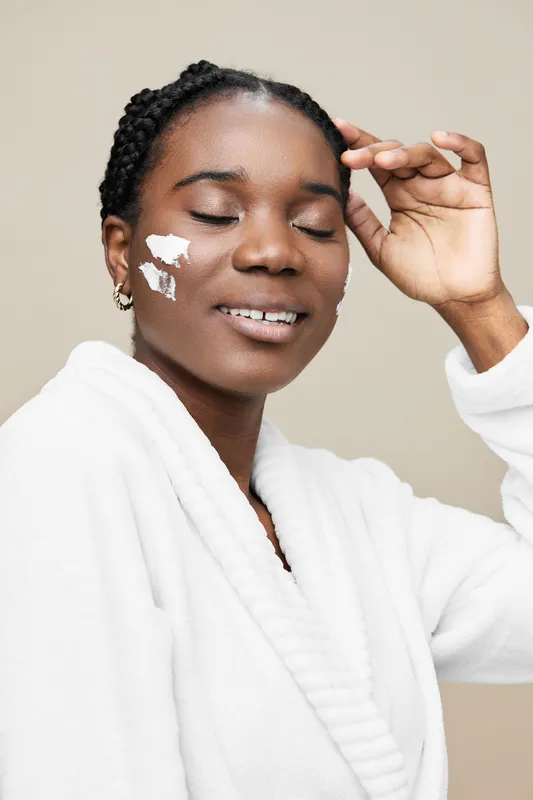 Young Black woman in a white robe applying skincare product, smiling with closed eyes, showcasing a peaceful and confident expression, light beige background