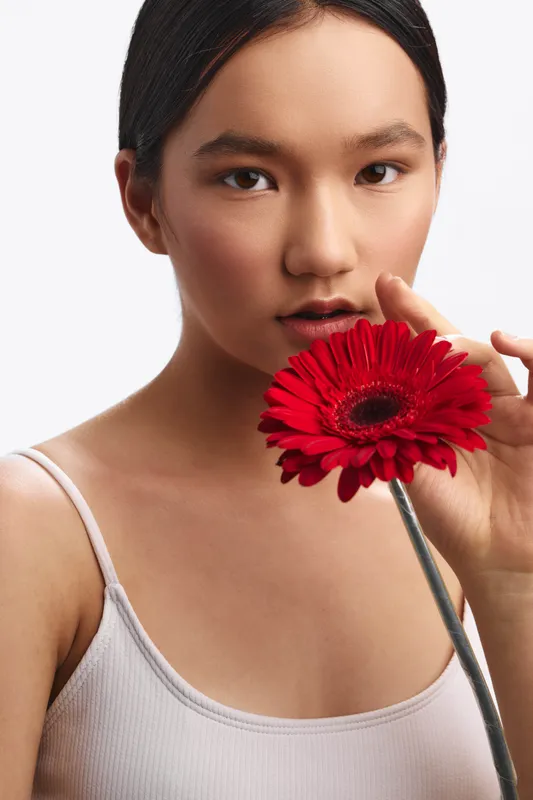 asian woman presents a graceful portrait as she holds a vibrant red flower near her lips, exuding calmness, elegance, and a thoughtful, intimate mood in a simple studio setting.