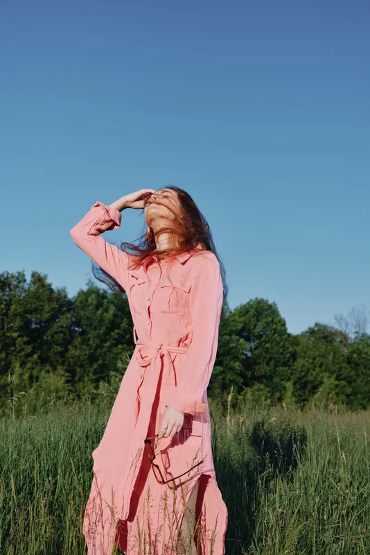 A woman in a flowing pink dress shielding her eyes from the sun, surrounded by vibrant green grass under a clear blue sky, exuding confidence and serenity