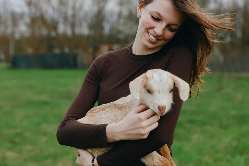Young woman in a brown sweater gently holds a baby goat outdoors on a cloudy day while smiling softly and enjoying the peaceful moment in a natural green field.