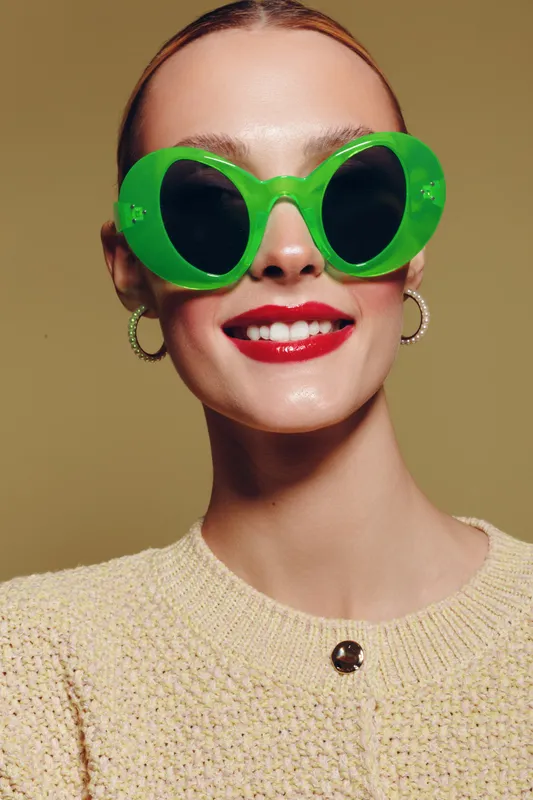 Close-up portrait of a smiling woman wearing large green sunglasses and red lipstick. She has hoop earrings and a beige knitted sweater against a neutral background. Fashion concept.