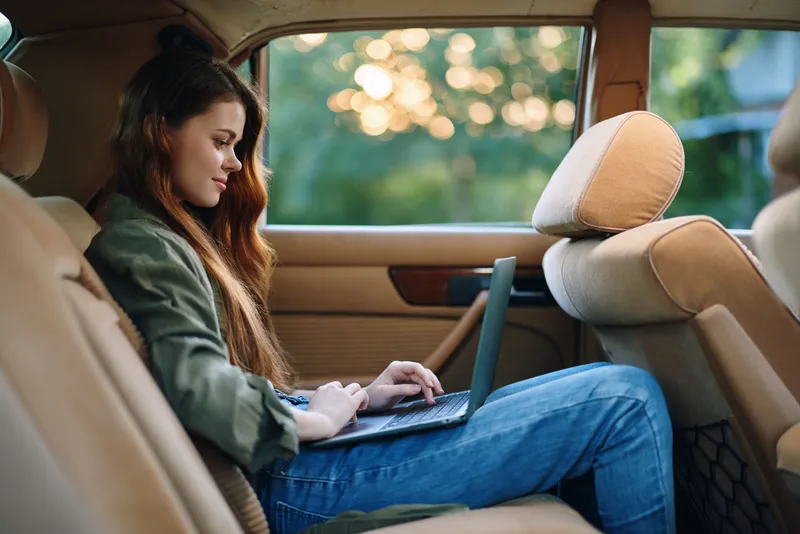 Young woman working on a laptop inside a car during sunset, showcasing a blend of productivity and relaxation with a serene background.