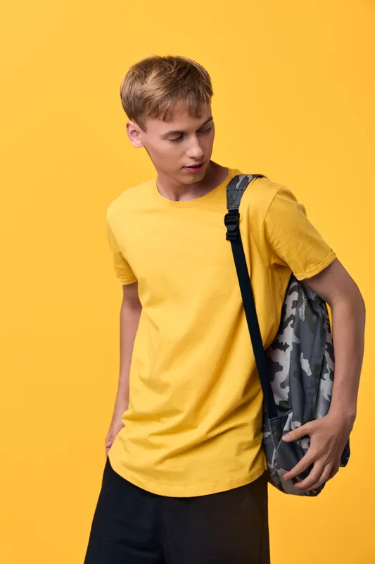 Man wearing a bright yellow shirt with a camouflage backpack on shoulder stands against a vivid yellow studio backdrop, conveying casual style, youth energy, and ready for activity