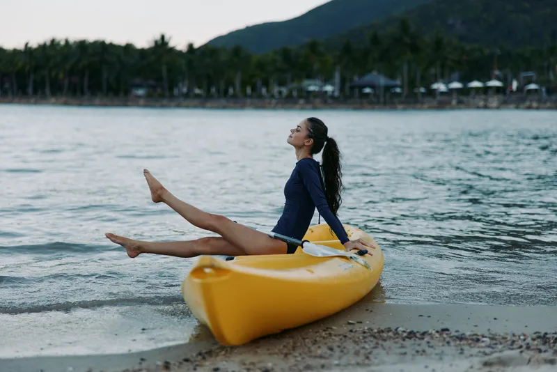 Relaxed woman sitting on a yellow kayak at the beach with mountains and palm trees in the background, enjoying calm water and outdoor leisure moments.
