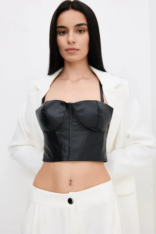 Young woman with long dark hair wearing black leather bustier and white pants stands confidently against plain background, showcasing modern fashion style and elegance.