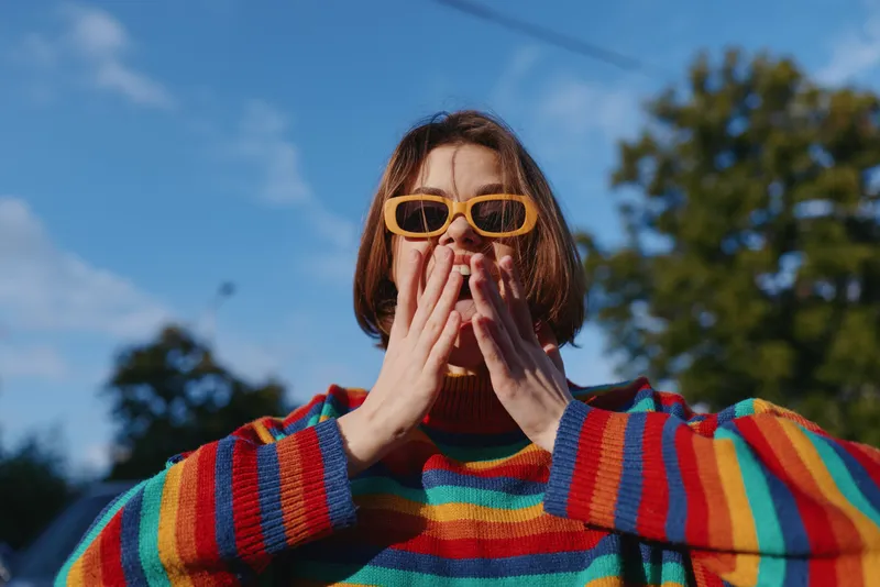 Young woman with short hair wearing a rainbow sweater and yellow sunglasses, smiling and playful outdoors in an urban park, candid fashion portrait radiating joyful mood.