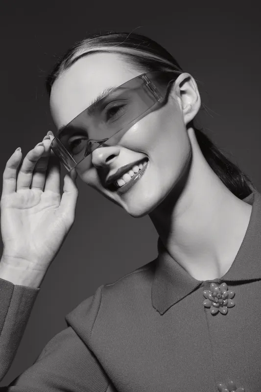 Young Caucasian woman with fair skin smiles wearing transparent sunglasses and a textured sweater with floral buttons on a dark background. Concept of fashion and style.