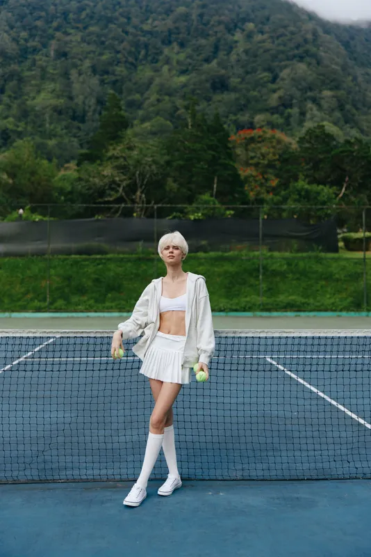 Fashionable young Caucasian woman in sporty outfit poses confidently on tennis court, showcasing vibrant colors and summer vibes while holding tennis balls