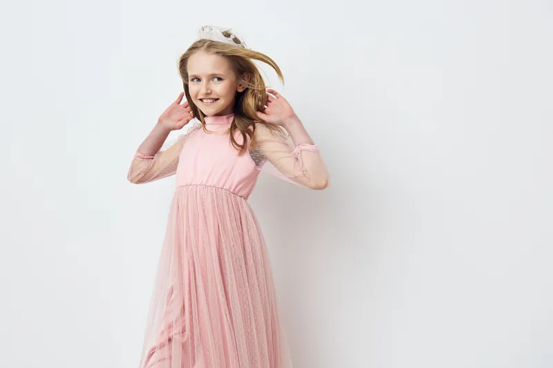 A cheerful girl in a light pink dress joyfully plays with her hair against a soft white background, showcasing childhood innocence and happiness