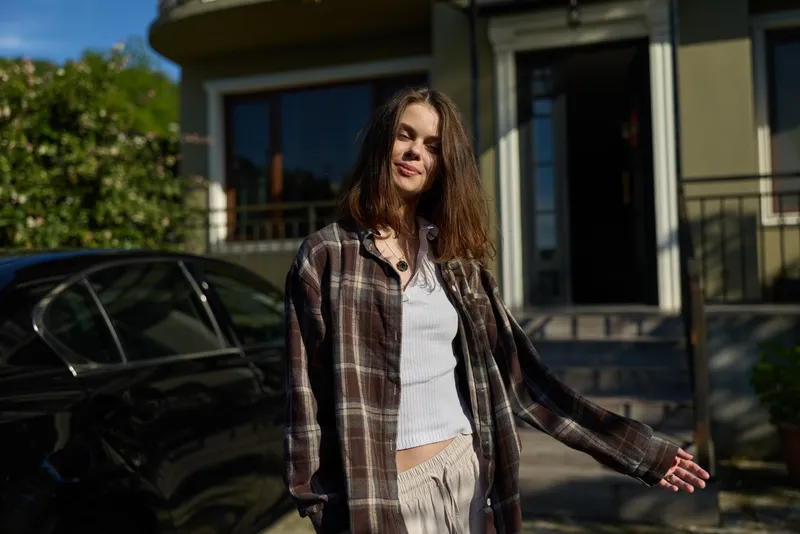 Casual woman lifestyle outdoor portrait in sunny daylight, wearing plaid shirt and white top, smiling near a modern home, conveying everyday relaxed mood