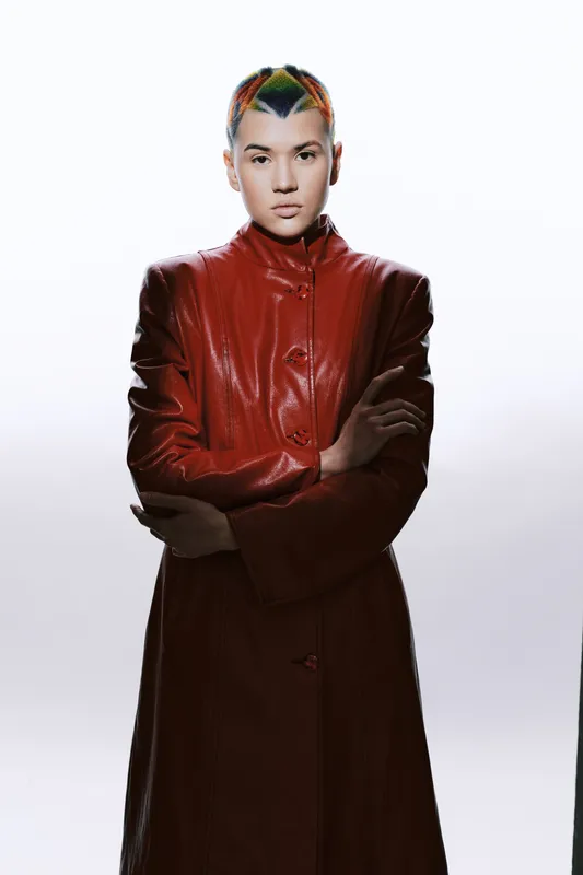 Young woman with short colorful hair wearing a red leather coat stands confidently against a bright background, exuding bold style and modern fashion attitude.