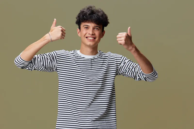 smiling man showing thumbs up wearing striped long sleeve shirt, curly hair, casual style, positive gesture against neutral background