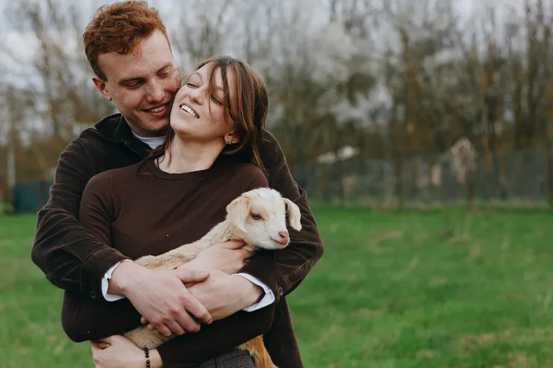 Joyful couple outdoors with a gentle goat kid, embracing and smiling, in a park setting, showcasing affectionate lifestyle moment, warm connection, and playful pet interaction