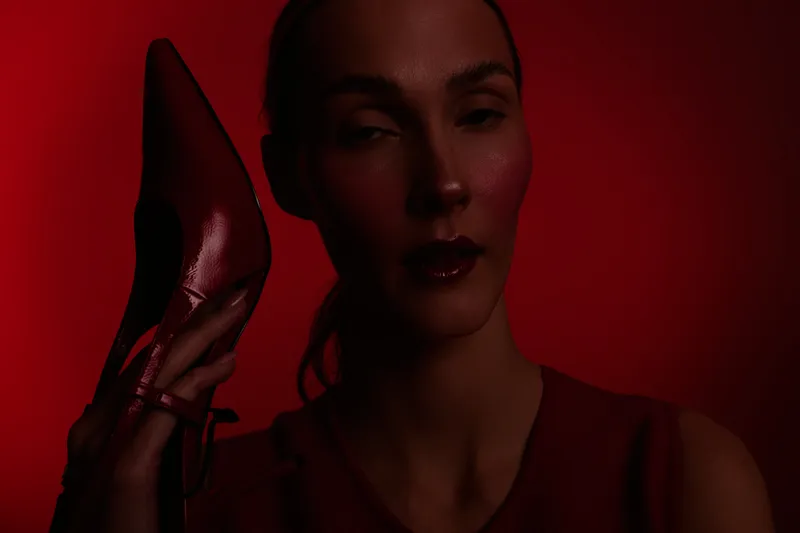 Portrait of a woman with dark makeup holding a red high heel shoe near her face against a red background. Concept of fashion, style, and elegance with dramatic lighting.