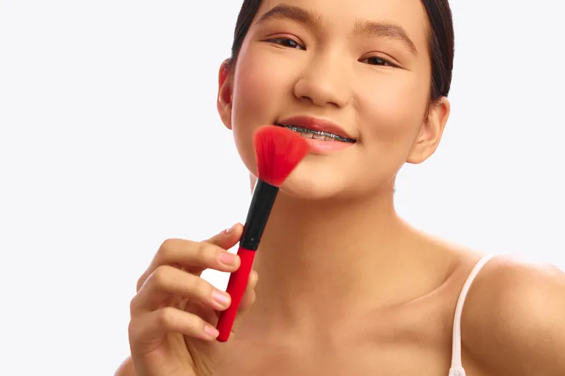 asian woman smiles softly while holding a makeup brush near her cheek in a bright studio. her clear skin and relaxed pose convey beauty, confidence, and everyday personal care.