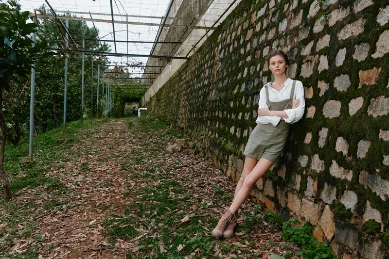 Young woman in green overalls stands confidently against a rustic stone wall in a lush garden, evoking a serene and natural vibe with earthy tones