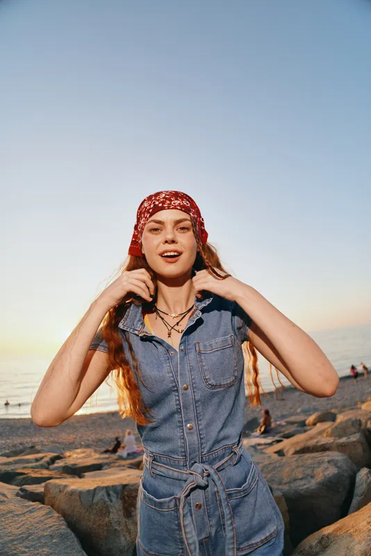 Lifestyle woman in modern boho-western denim street style wearing a red bandana by the beach at sunset with warm film color tones and relaxed natural vibe.